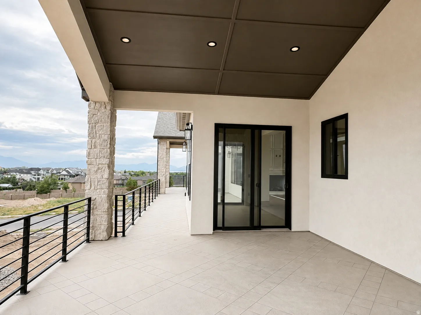 Expansive covered balcony featuring stone-clad pillars, black metal railings, recessed lighting, and large sliding glass doors