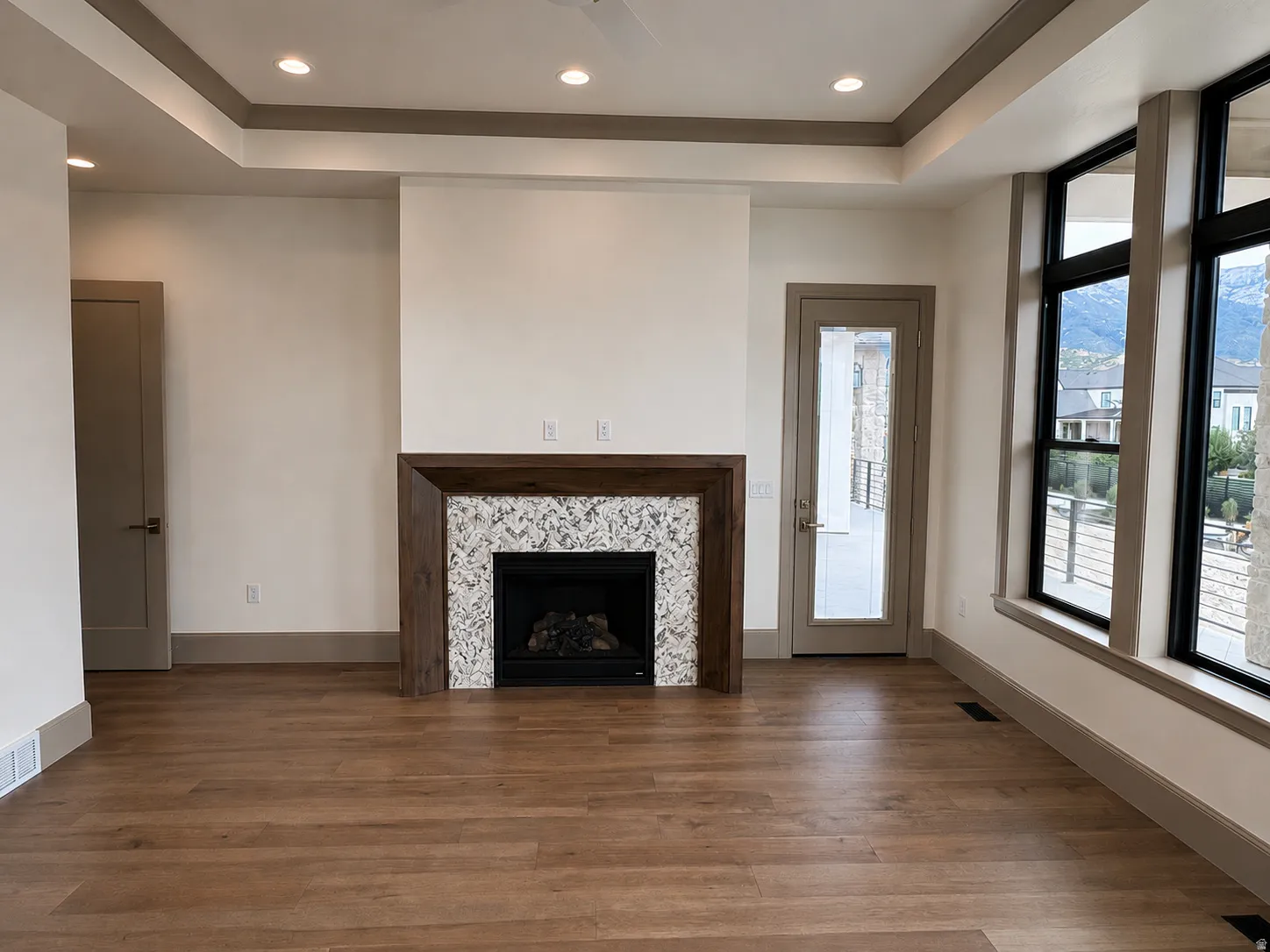 Recessed lighting with a tray ceiling, wood-finish flooring, built-in fireplace with a stone surround and wood mantle, and large windows with black frames