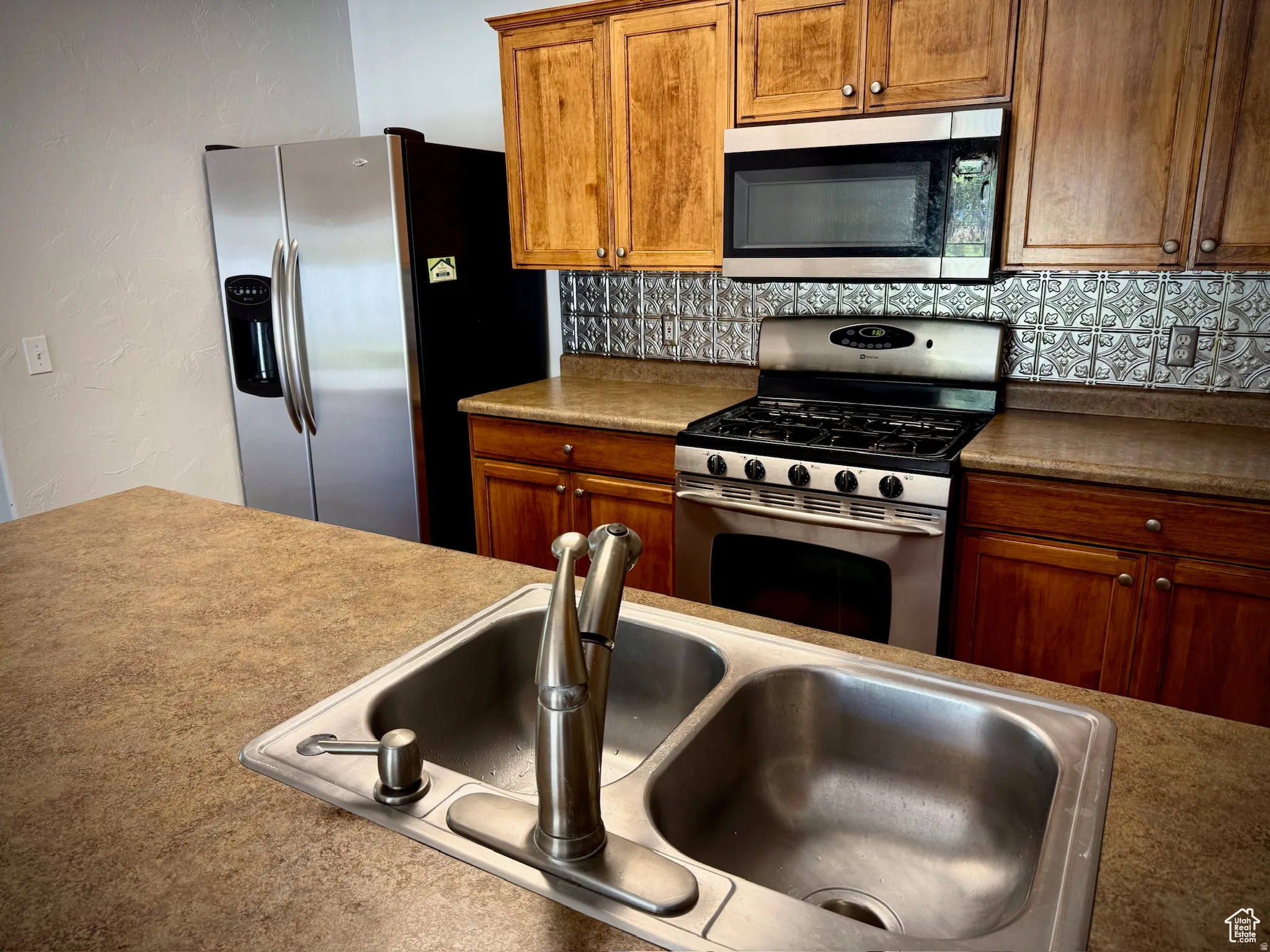 Kitchen with stainless steel appliances, a textured wall, wood finish cabinets, and tasteful backsplash