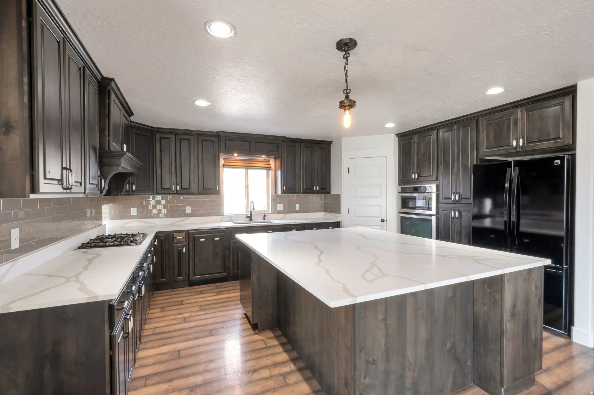 Spacious kitchen featuring an expansive island with a white veined countertop, dark wood-finish cabinetry, a built-in gas cooktop, and a tile backsplash