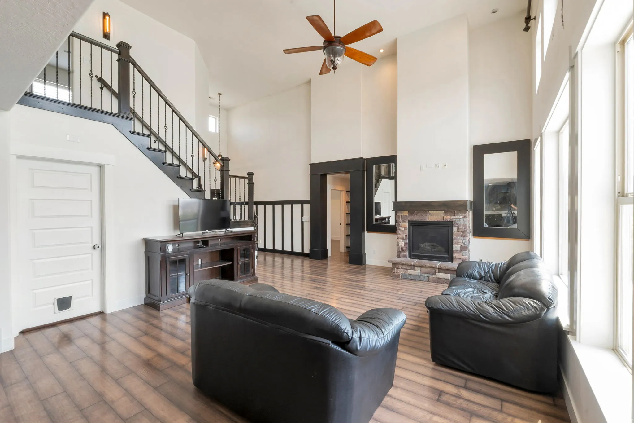 Spacious room featuring high ceilings, wood-finish flooring, a stone fireplace, large windows, and a staircase with wrought iron balusters