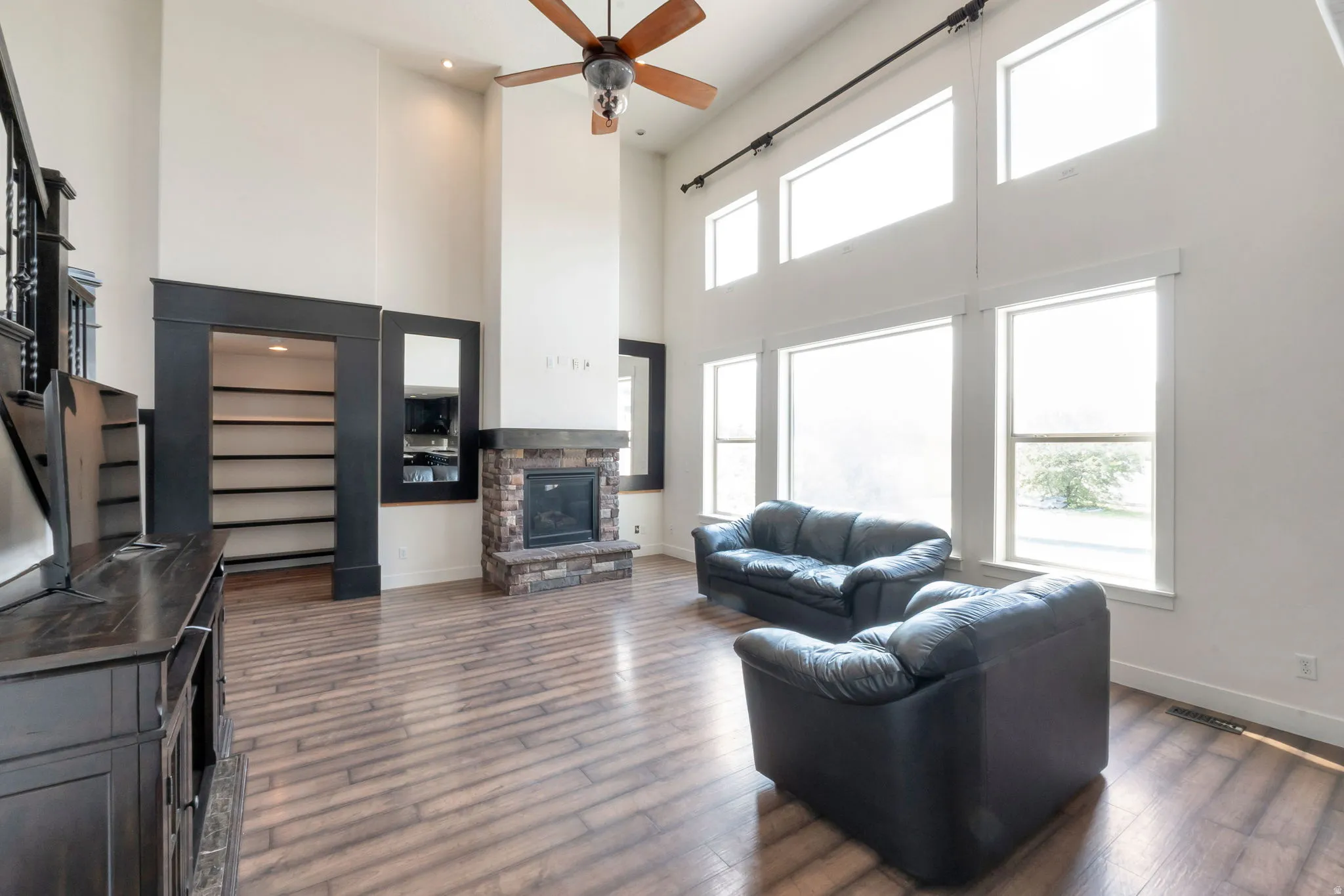 Vaulted ceiling great room featuring wood-finish flooring, a stone-surround fireplace with hearth, and extensive natural light through a wall of windows