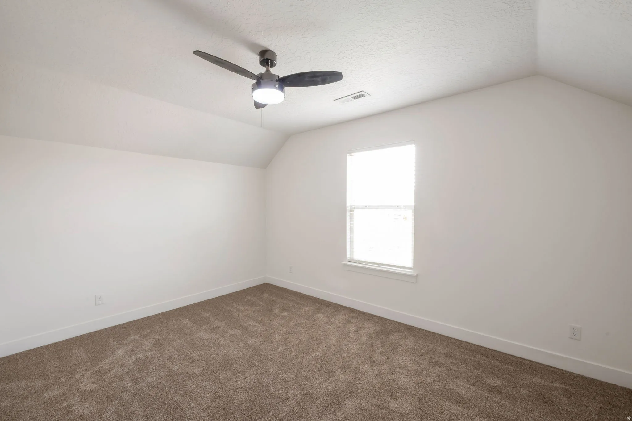 Spacious room featuring vaulted ceilings, a single window, a modern ceiling fan with integrated lighting, and plush brown carpeting
