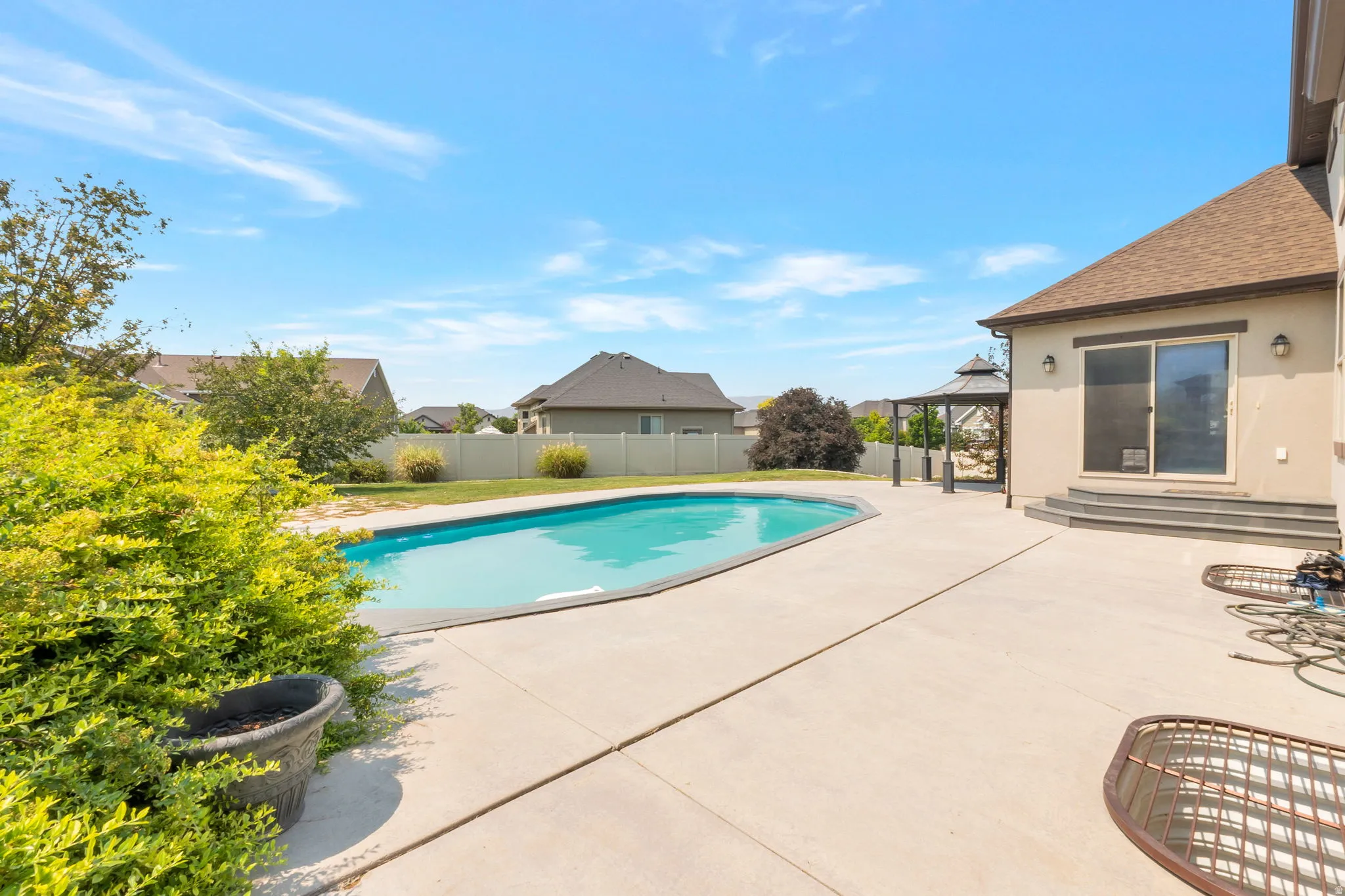 Expansive concrete patio featuring a curved inground swimming pool, a covered gazebo, and direct access to the interior via a sliding glass door