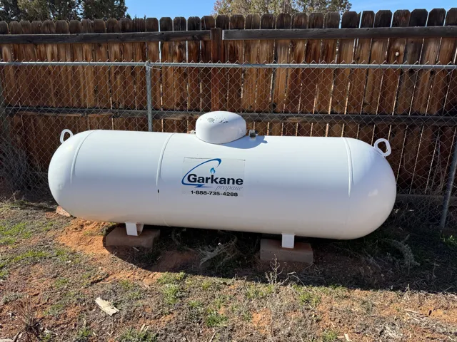 Exterior propane tank with a white finish, supported by concrete blocks, situated against a mixed wood and chain-link fence