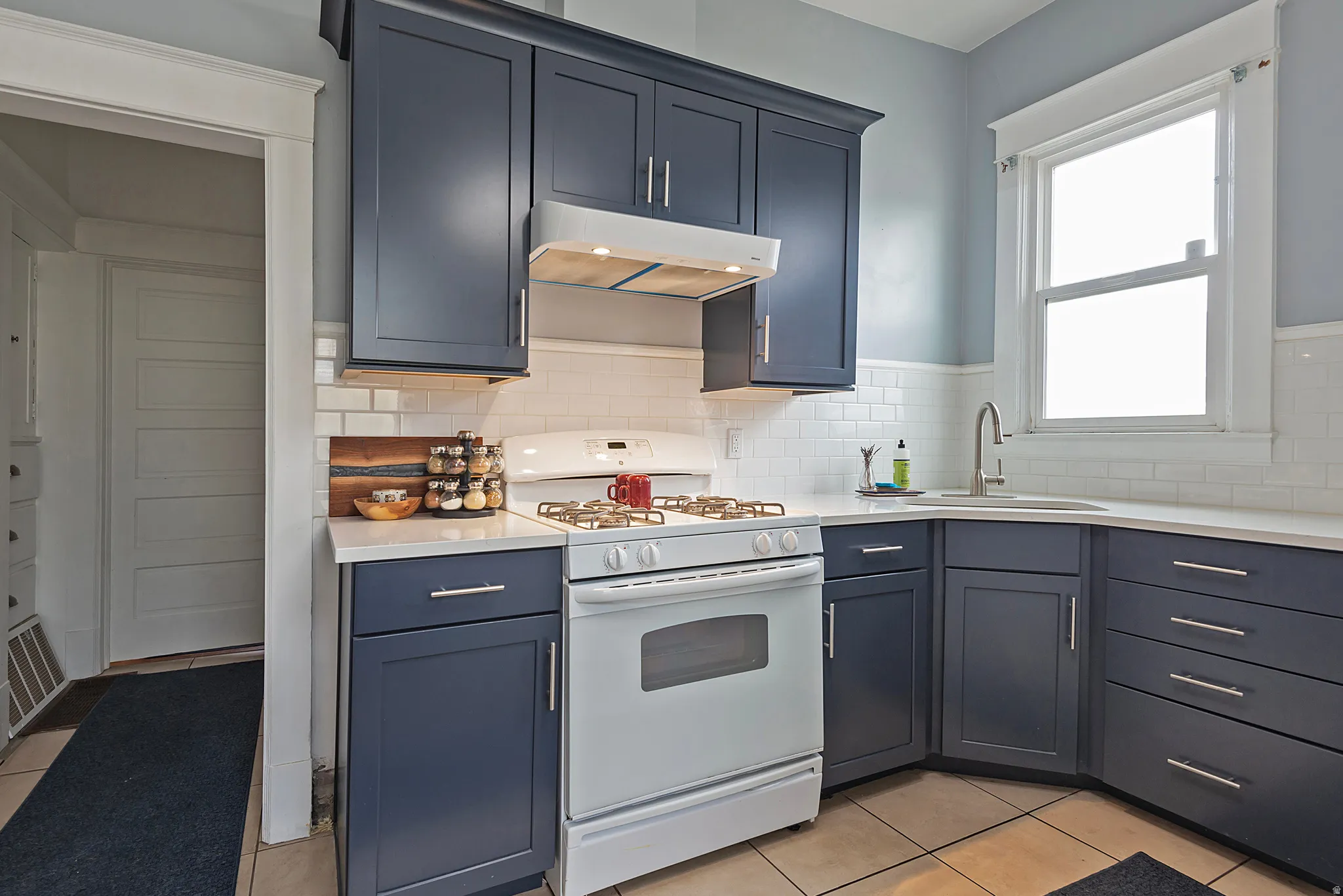 Kitchen featuring white range with gas stovetop, light tile patterned floors, light stone countertops, backsplash, and blue cabinetry