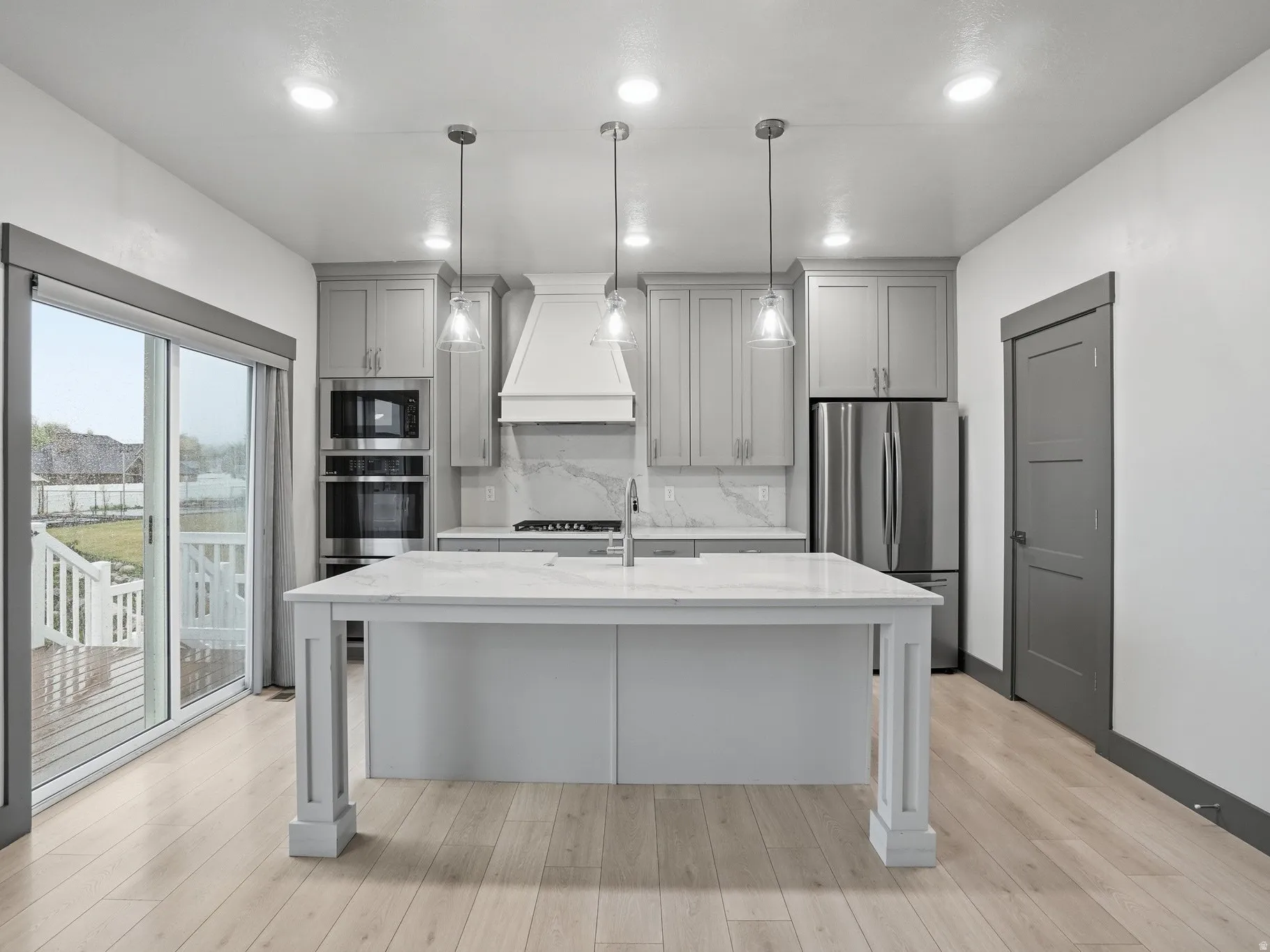 Kitchen with gray cabinetry, pendant lighting, light stone counters, stainless steel appliances, and a center island with sink