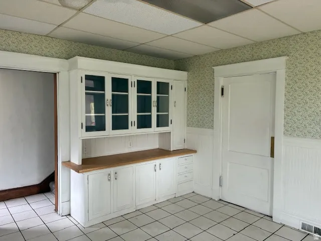 Kitchen featuring wainscoting, a drop ceiling, white cabinetry, wallpapered walls, and light tile patterned floors