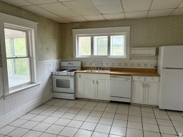 Kitchen featuring white appliances, wallpapered walls, a drop ceiling, white cabinetry, and light tile patterned flooring