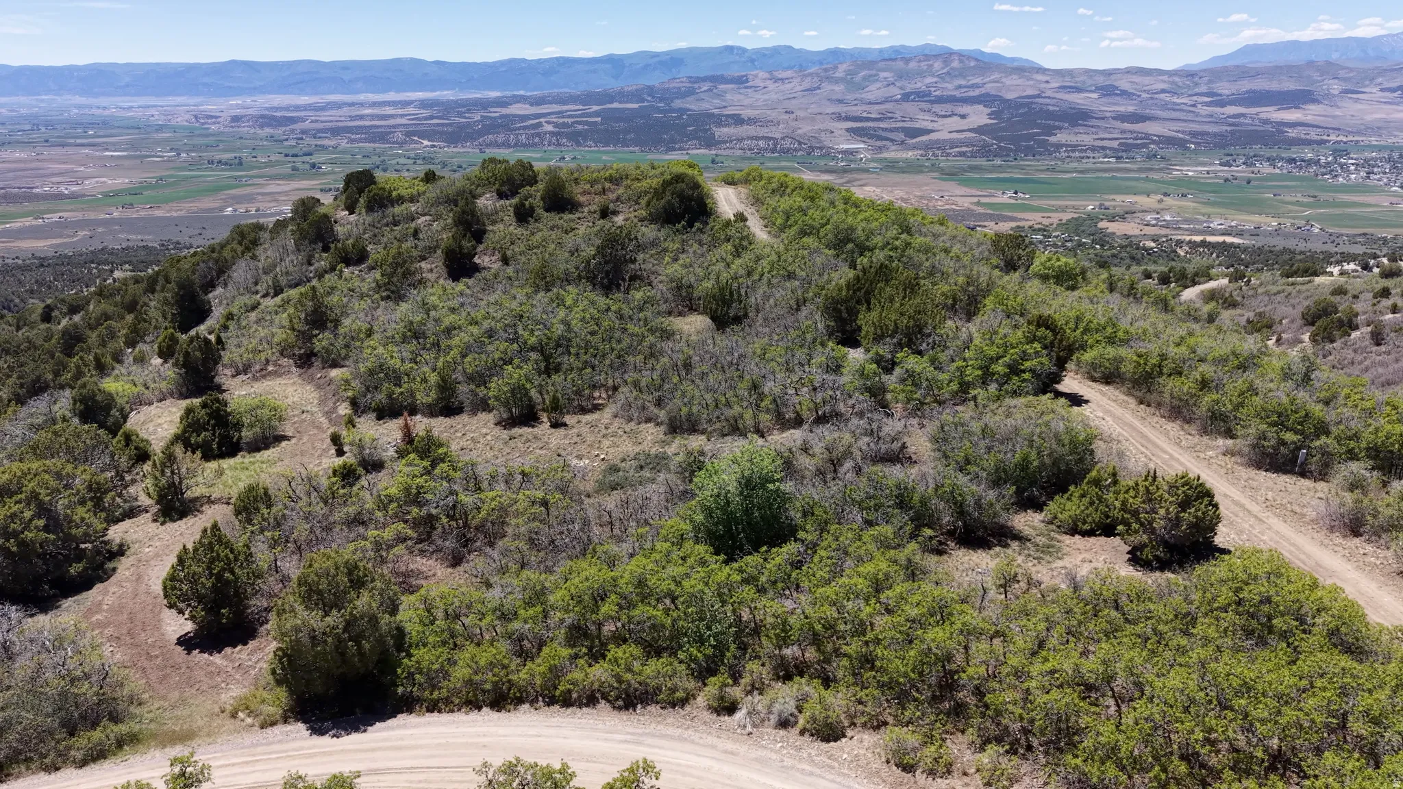 Expansive mountain terrain featuring natural vegetation, multiple dirt paths, and distant valley and mountain ranges under a clear sky