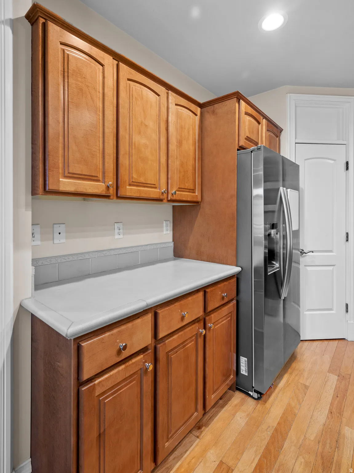 Kitchen featuring light countertops, stainless steel refrigerator with ice dispenser, light wood-type flooring, wood finish cabinetry, and recessed lighting