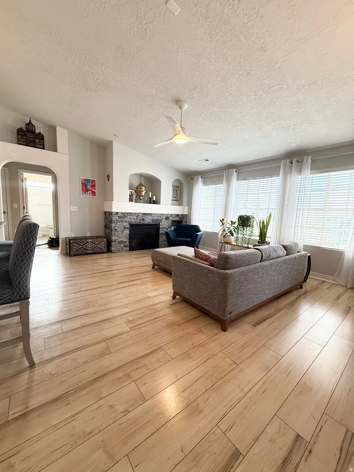 Living room featuring a stone fireplace, ceiling fan, a textured ceiling, arched walkways, and light wood finished floors