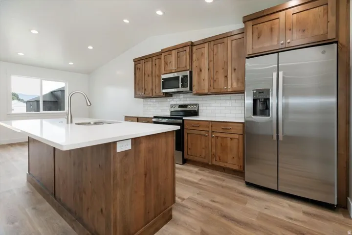 Kitchen featuring vaulted ceiling, stainless steel appliances, decorative backsplash, light wood finished floors, and a center island with sink
