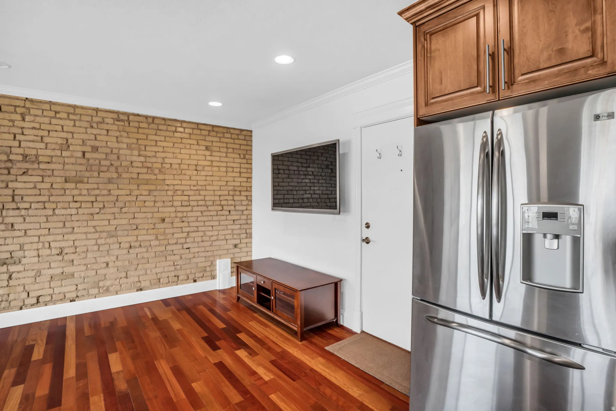 Kitchen with brick wall, stainless steel refrigerator with ice dispenser, crown molding, dark wood-type flooring, and recessed lighting