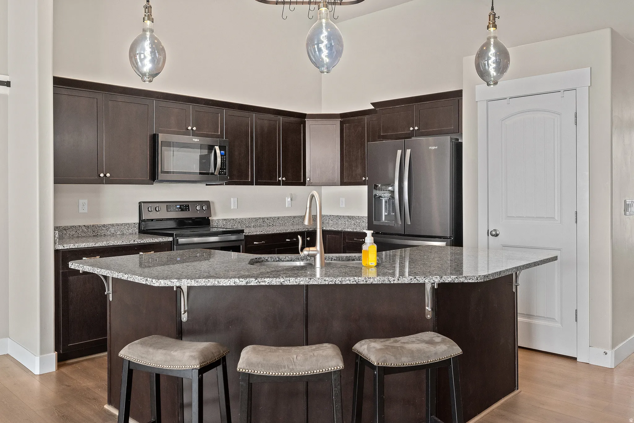 Kitchen featuring dark wood finish cabinetry, stainless steel appliances, light stone counters, a center island with sink, and pendant lighting