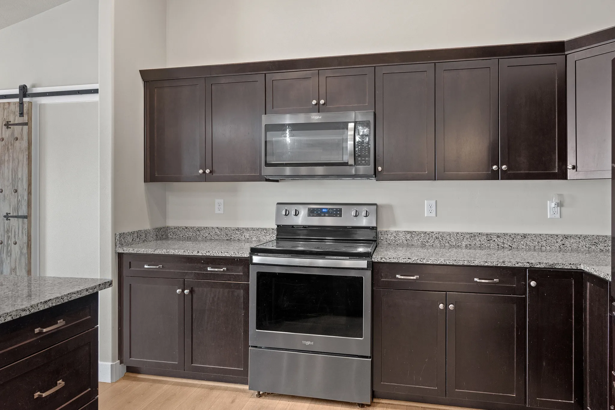 Kitchen featuring dark wood finish cabinets, stainless steel appliances, light stone counters, a barn door, and light wood finished floors