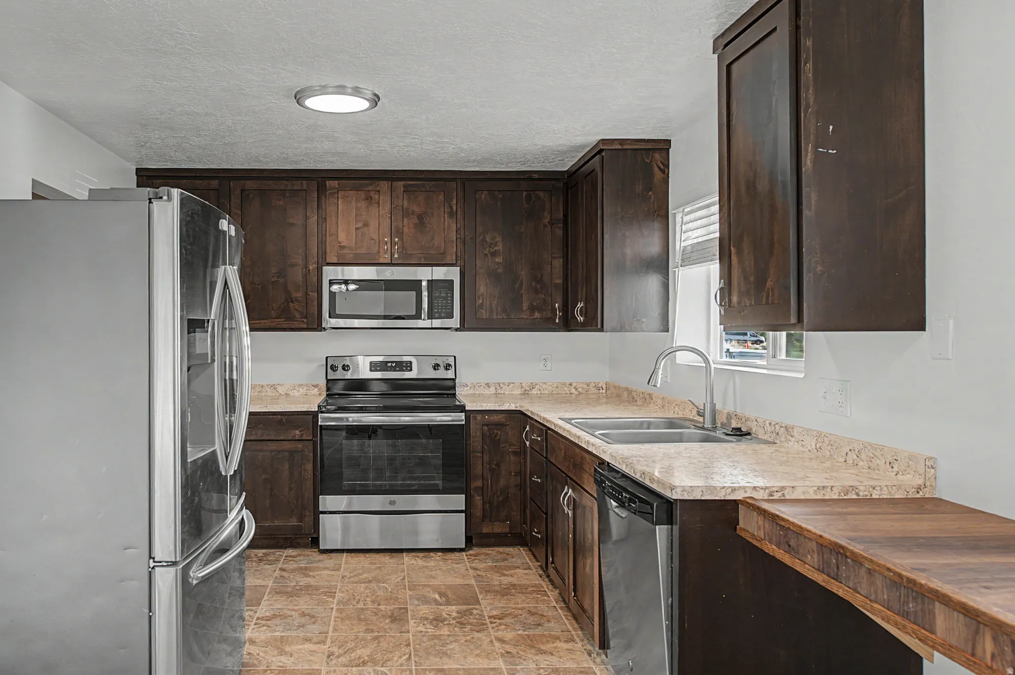 Kitchen featuring dark wood finish cabinets, stainless steel appliances, stone finish flooring, and a textured ceiling