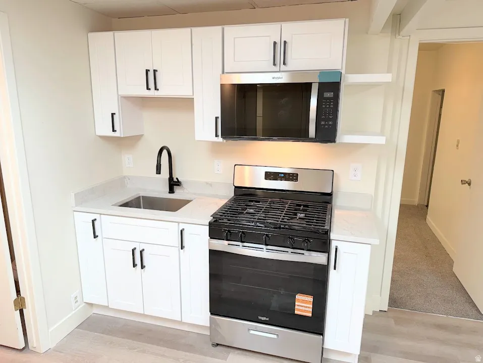 Compact kitchen featuring white shaker cabinetry, stainless steel appliances, light-toned countertops, a black gooseneck faucet, and wood-finish flooring