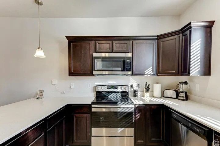 Kitchen featuring stainless steel appliances, dark wood finish cabinets, decorative light fixtures, and light stone countertops