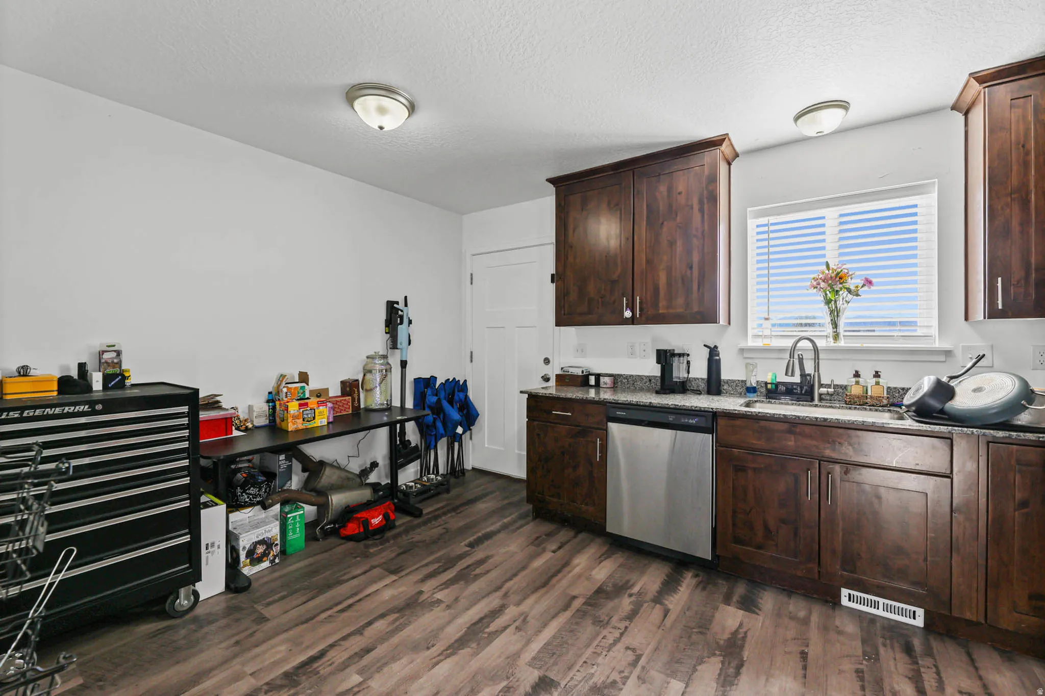 Kitchen featuring dark wood finish cabinets, stainless steel dishwasher, dark wood-style flooring, a textured ceiling, and light stone counters