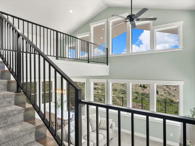 Two-story living area featuring a vaulted ceiling with a ceiling fan, multiple large windows, and a carpeted staircase with dark metal railings