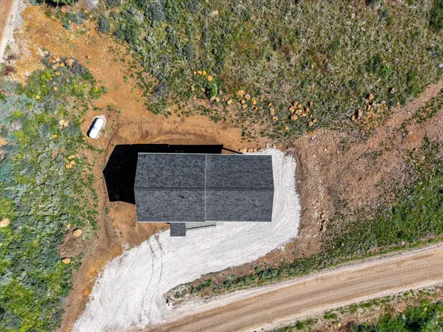 New construction featuring a dark shingle roof, surrounded by natural terrain and a gravel driveway