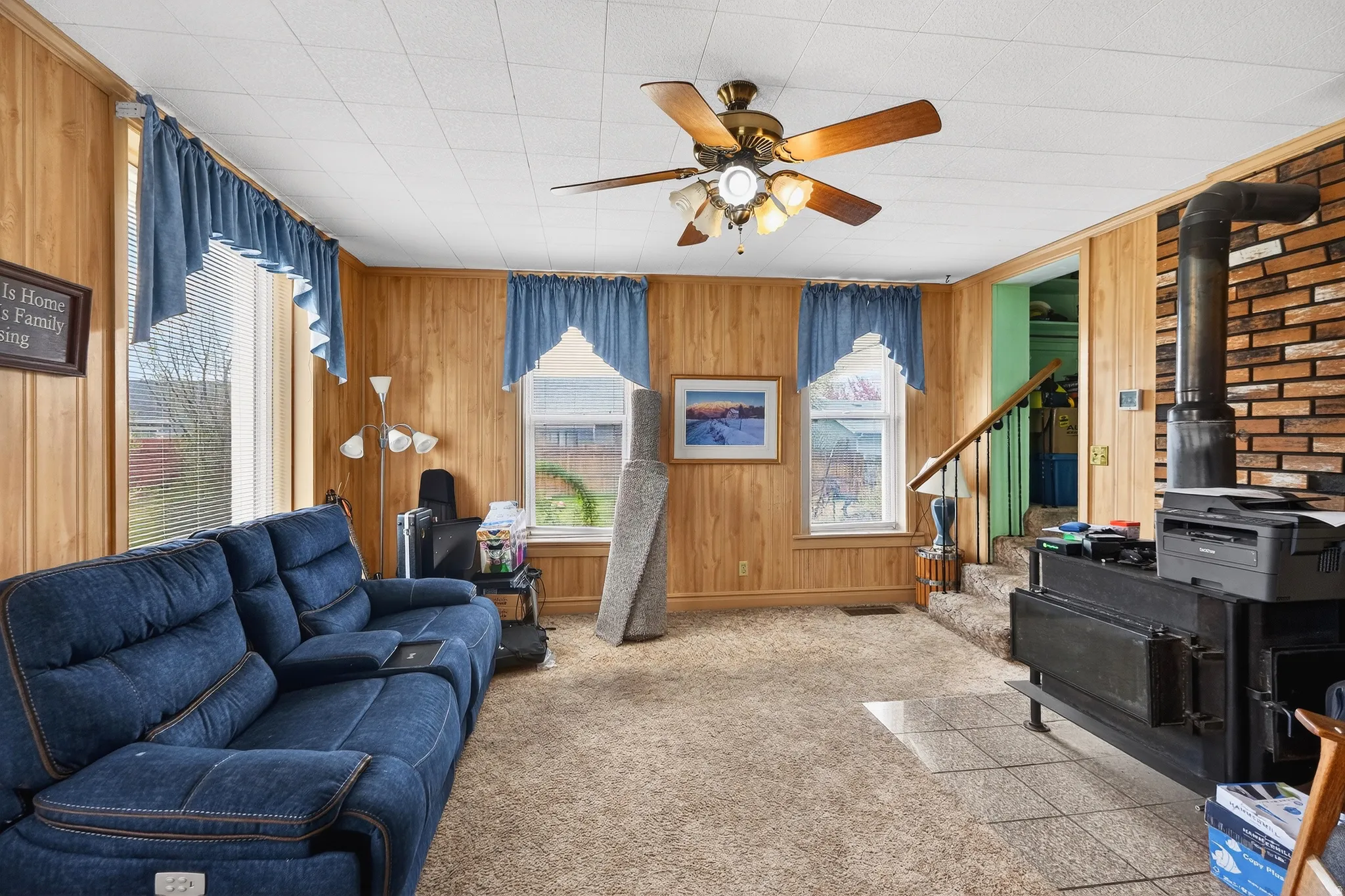 Living area with a wood stove, ceiling fan, light colored carpet, and wood walls