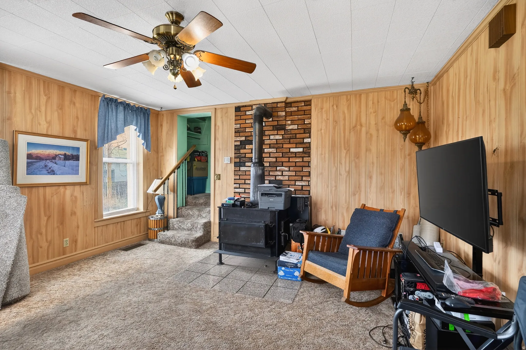 Living area featuring wood walls, a wood stove, light colored carpet, and a ceiling fan