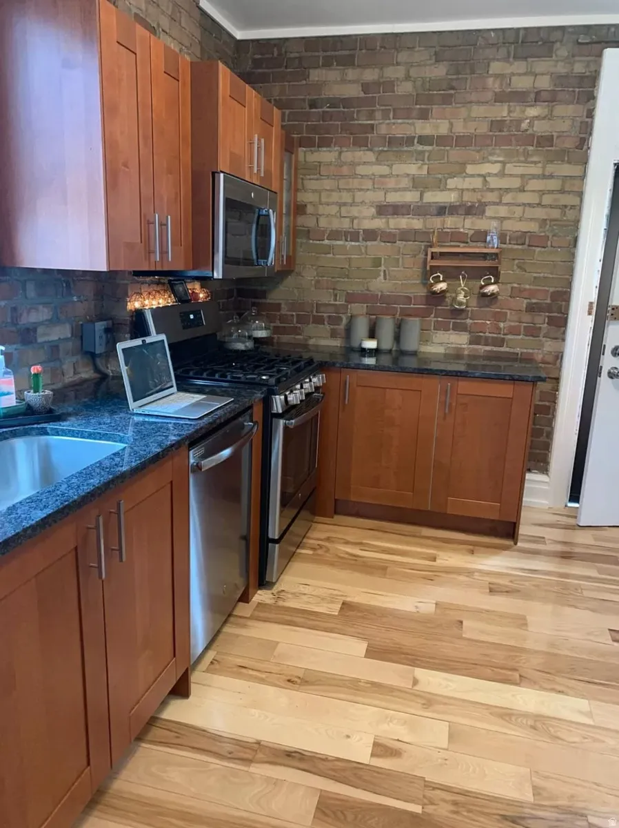 Kitchen featuring wood finish cabinets, stainless steel appliances, dark stone countertops, light wood-style flooring, and brick wall