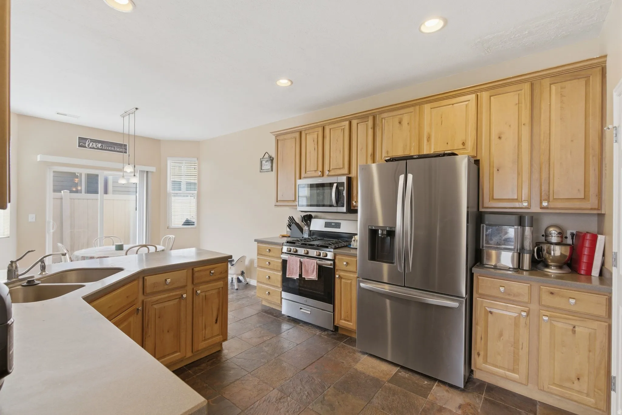 Kitchen with stainless steel appliances, stone tile flooring, decorative light fixtures, and light wood finish cabinets