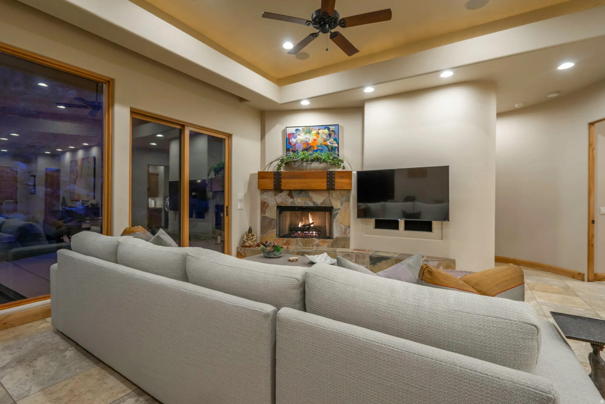 Living room with stone tile flooring, a ceiling fan, recessed lighting, an outdoor stone fireplace, and a tray ceiling