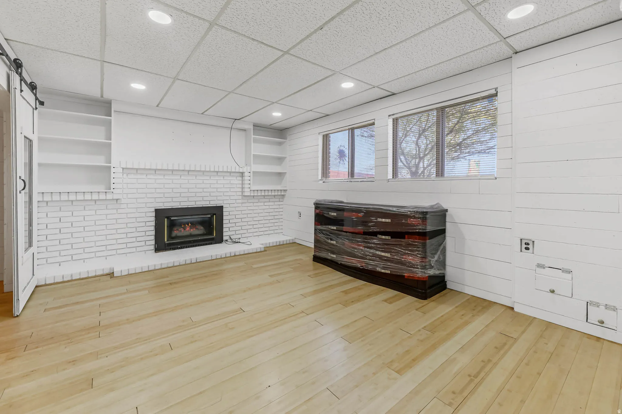 Unfurnished living room with built in features, light wood-type flooring, a paneled ceiling, a barn door, and a fireplace