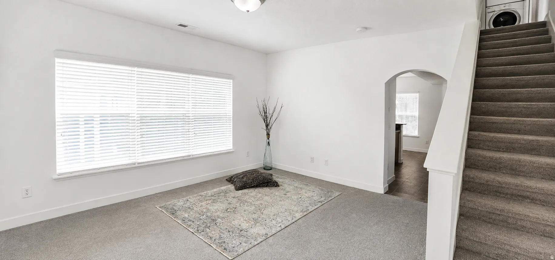 Carpeted living space with a large window featuring horizontal blinds, an arched doorway, and a carpeted staircase with a built-in washer and dryer unit