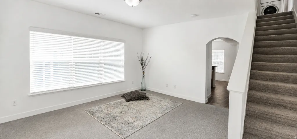 Carpeted living space with a large window featuring horizontal blinds, an arched doorway, and a carpeted staircase with a built-in washer and dryer unit