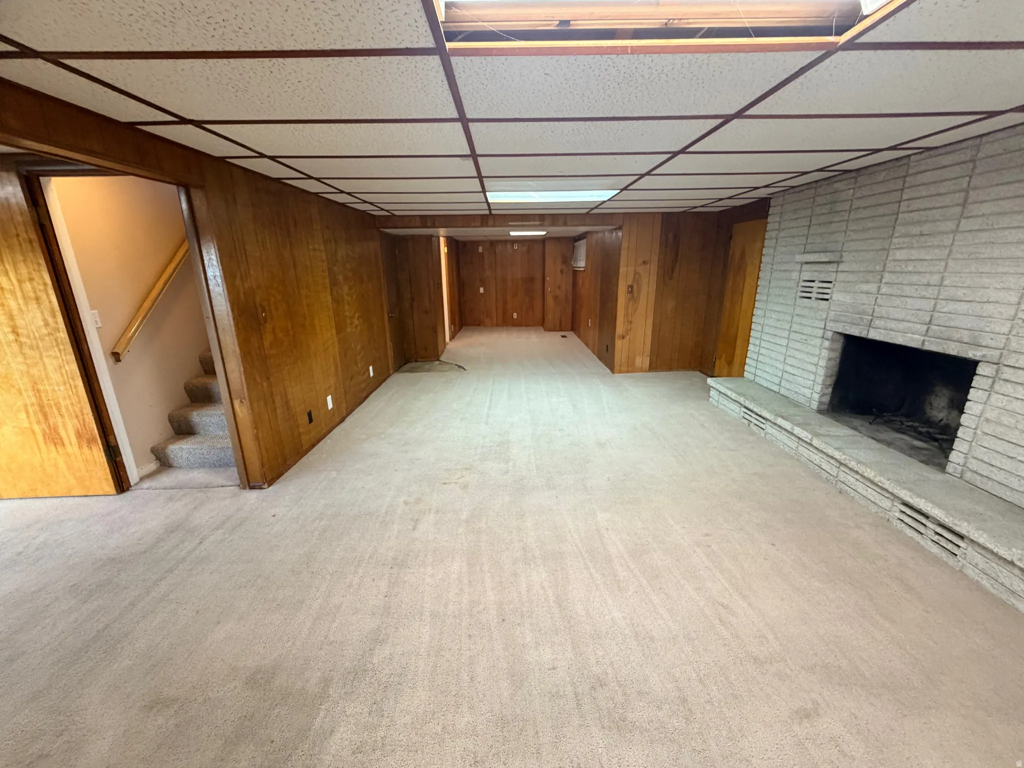Finished below grade area featuring light colored carpet, a brick fireplace, wood walls, and a paneled ceiling