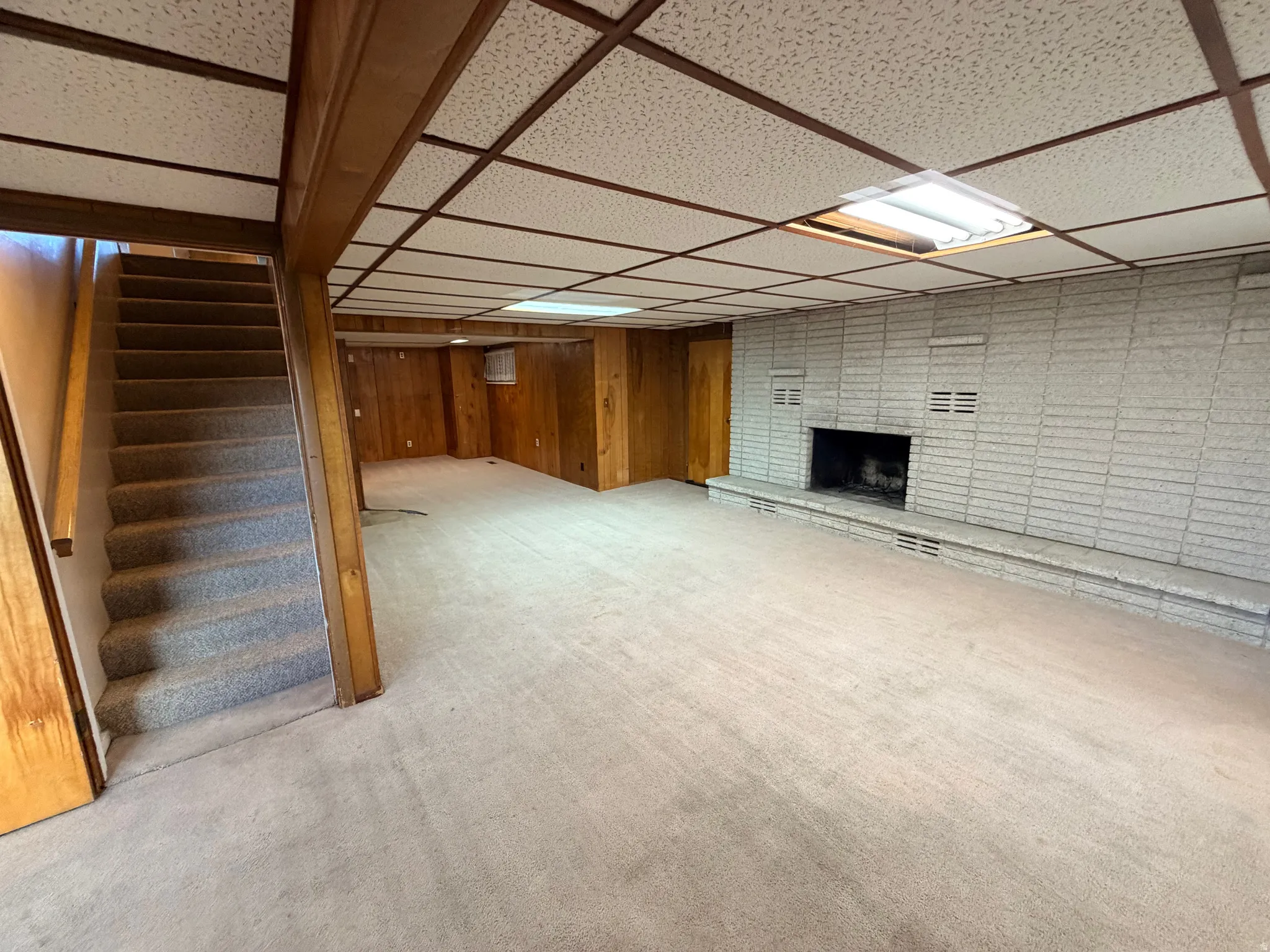 Finished basement with a brick fireplace, light colored carpet, a paneled ceiling, and wooden walls
