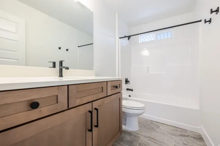 Full bathroom featuring a wood-finish vanity with a light-toned countertop, a black-finish faucet, a white tub-shower combination, and large format floor tiling