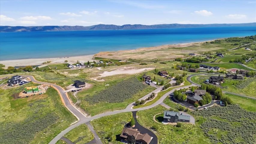 Expansive aerial perspective showcasing a residential community with paved roads, surrounded by lush green landscapes, with a tranquil lake and distant mountains forming a scenic backdrop