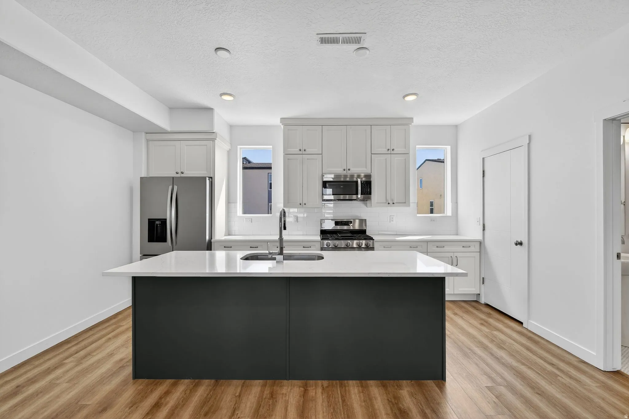 Kitchen with stainless steel appliances, light wood-type flooring, light stone countertops, a center island with sink, and backsplash