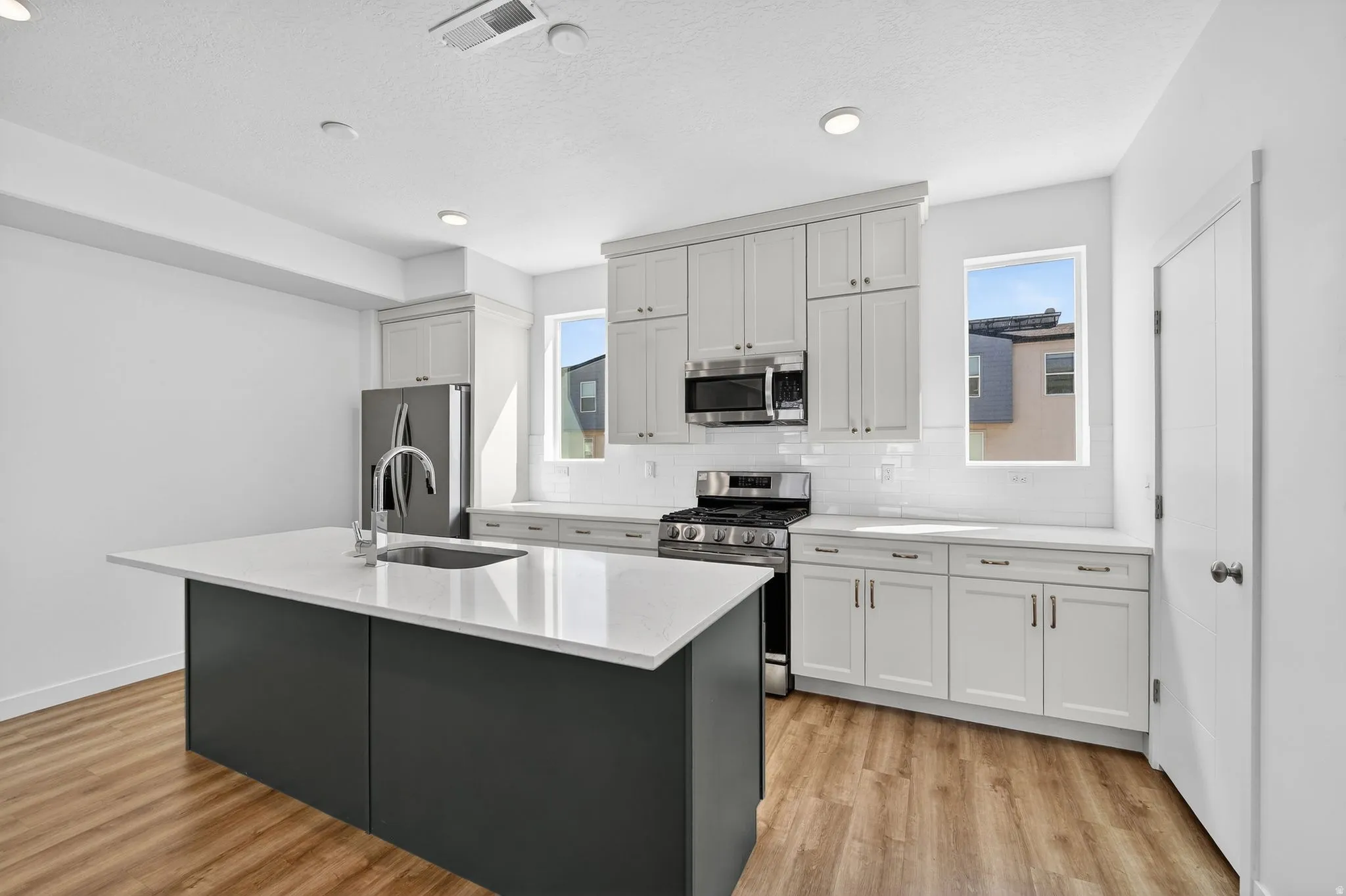 Kitchen featuring stainless steel appliances, light wood finished floors, decorative backsplash, light stone counters, and a kitchen island with sink
