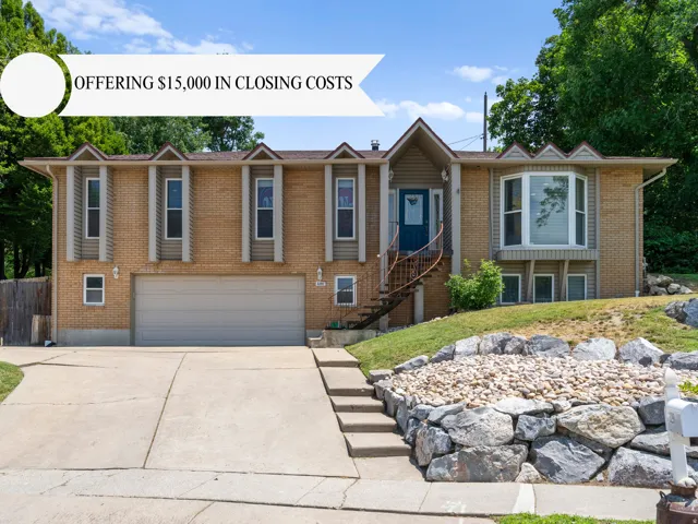 Split-level residence featuring a brick and siding exterior, a two-car garage, a concrete driveway, an external staircase with a curved metal railing, and a rock retaining wall with landscaping