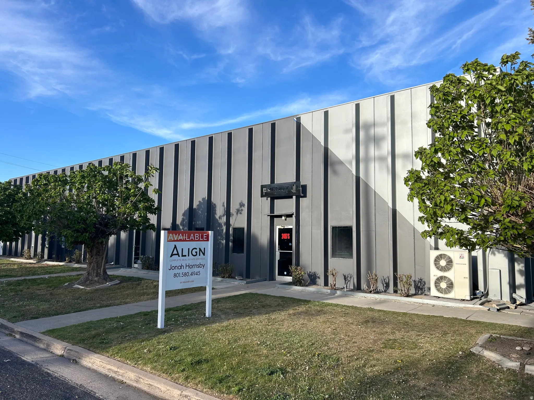 Commercial building featuring a grey facade with vertical accent panels, multiple windows, a prominent entrance, exterior HVAC units, and a paved walkway
