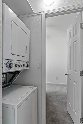 Laundry area featuring a textured wall, light carpet, stacked washing machine and dryer, and a textured ceiling