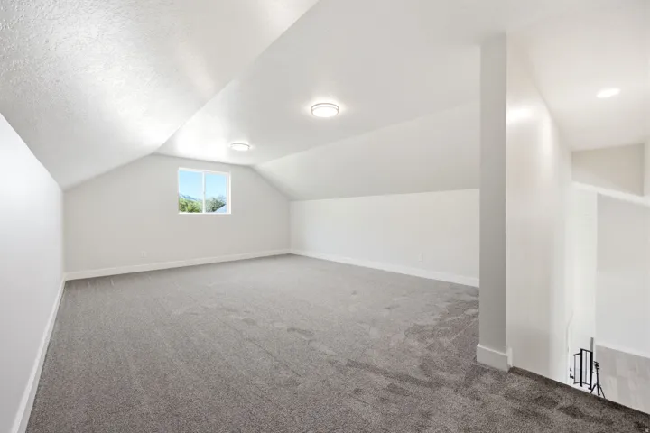 Spacious upper-level room featuring a window with exterior visibility, overhead recessed lighting, sloped ceiling architecture, and neutral-toned carpeting