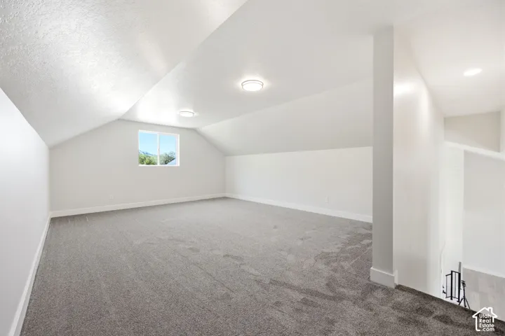 Spacious upper-level room featuring a window with exterior visibility, overhead recessed lighting, sloped ceiling architecture, and neutral-toned carpeting