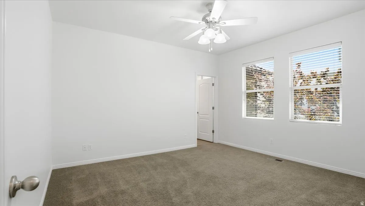 Spacious room featuring neutral carpeting, white painted walls, two windows with blinds, a white ceiling fan with light fixture, and a white interior door