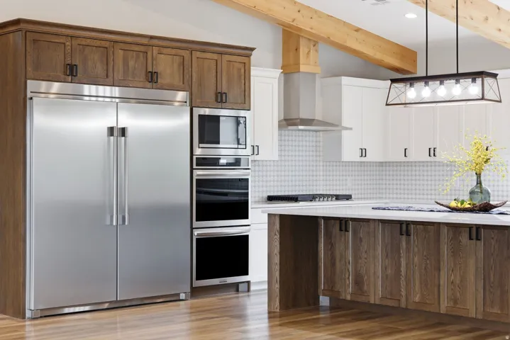 Two tone kitchen with built in appliances, decorative backsplash, pendant lighting, light stone countertops, and beam ceiling