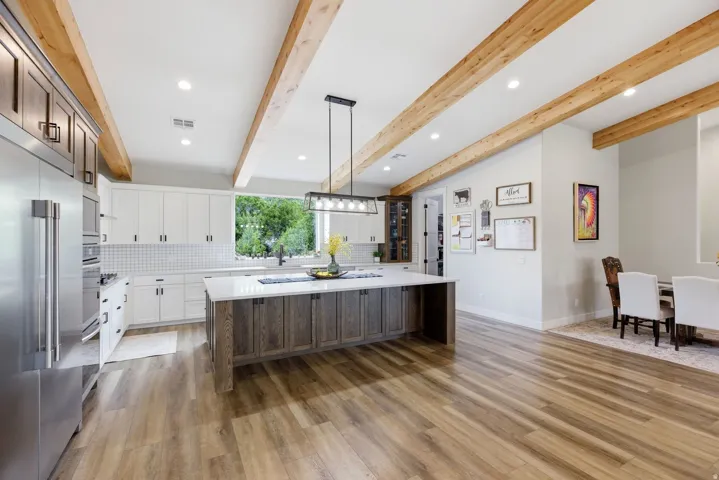 Two tone kitchen featuring a spacious island, stainless steel appliances, backsplash, light stone counters, and beam ceiling