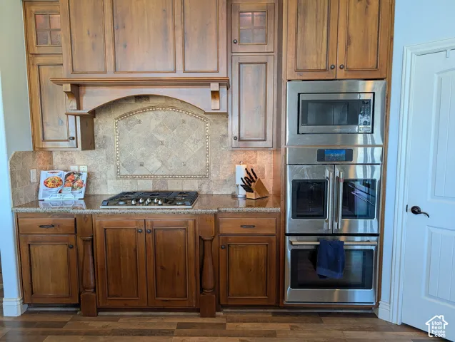 Kitchen with stainless steel appliances, dark stone counters, wood finish cabinetry, glass fronted cabinets, and tasteful backsplash