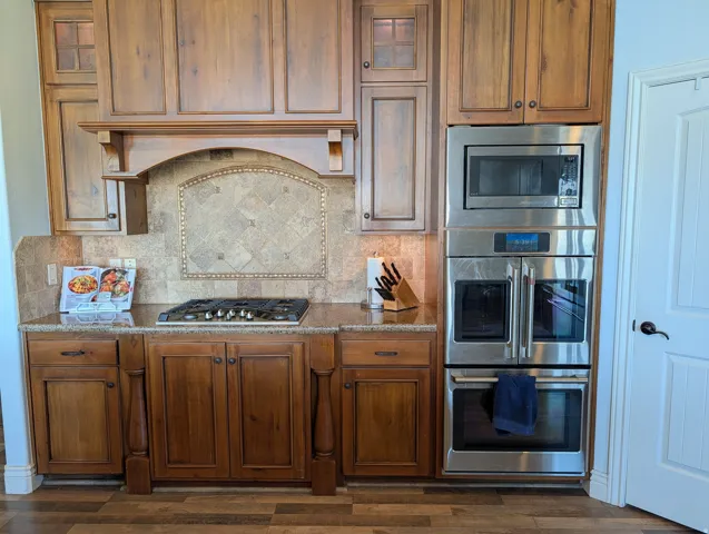 Kitchen with stainless steel appliances, dark stone counters, wood finish cabinetry, glass fronted cabinets, and tasteful backsplash